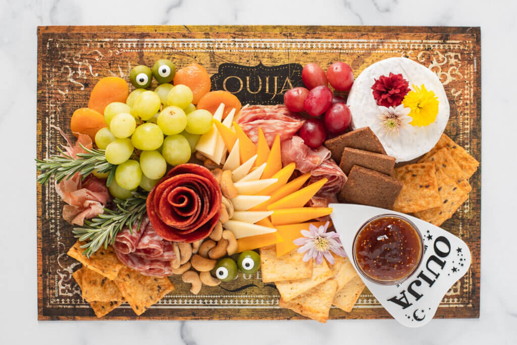 Halloween charcuterie board with a pepperoni rose, orange and white cheese triangles, cured meats, crackers, grapes, olives with candy eyes, Brie, cashews, and jam and a mum and rosemary garnish.
