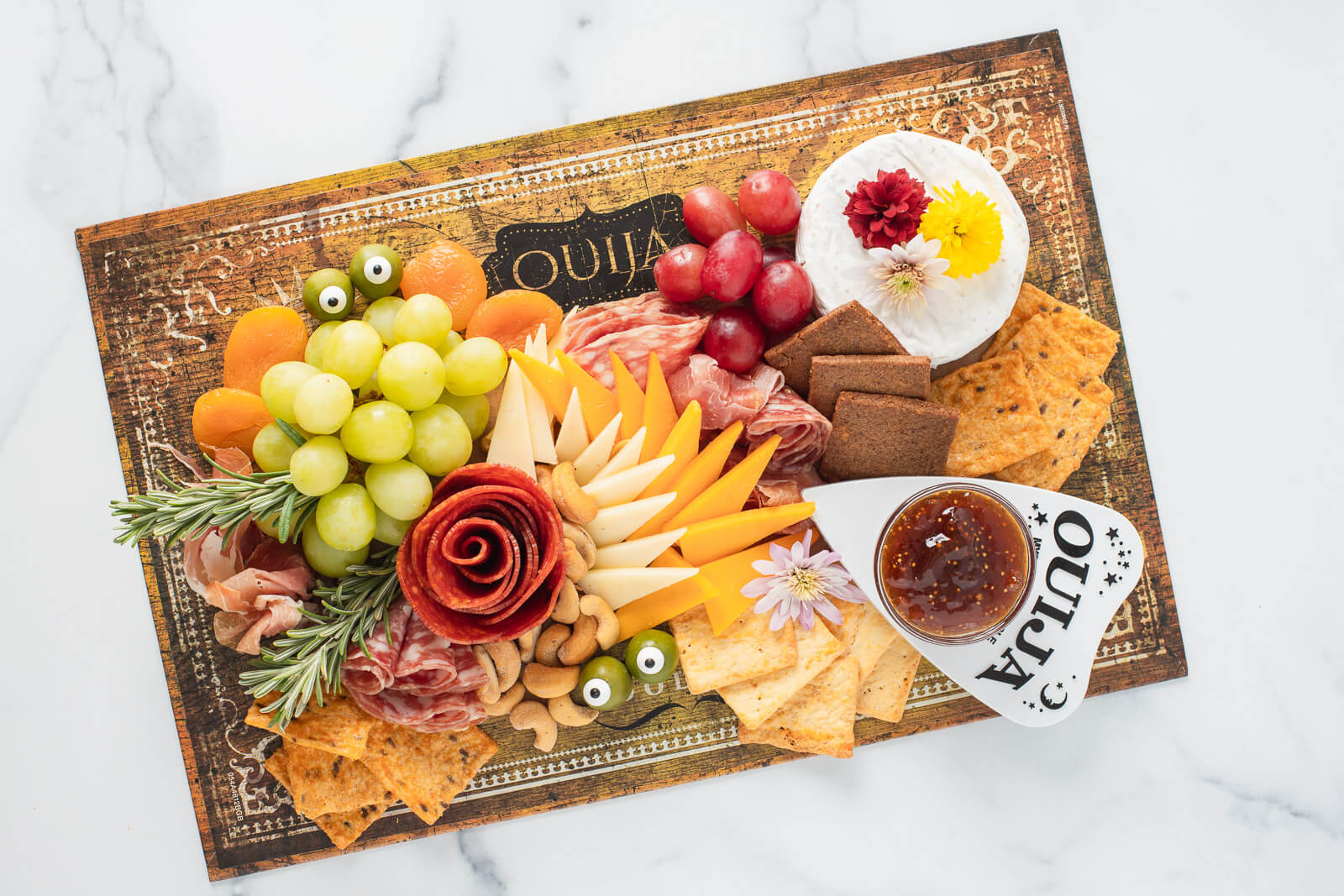Halloween charcuterie board with a pepperoni rose, orange and white cheese triangles, cured meats, crackers, grapes, olives with candy eyes, Brie, cashews, and jam and a mum and rosemary garnish.